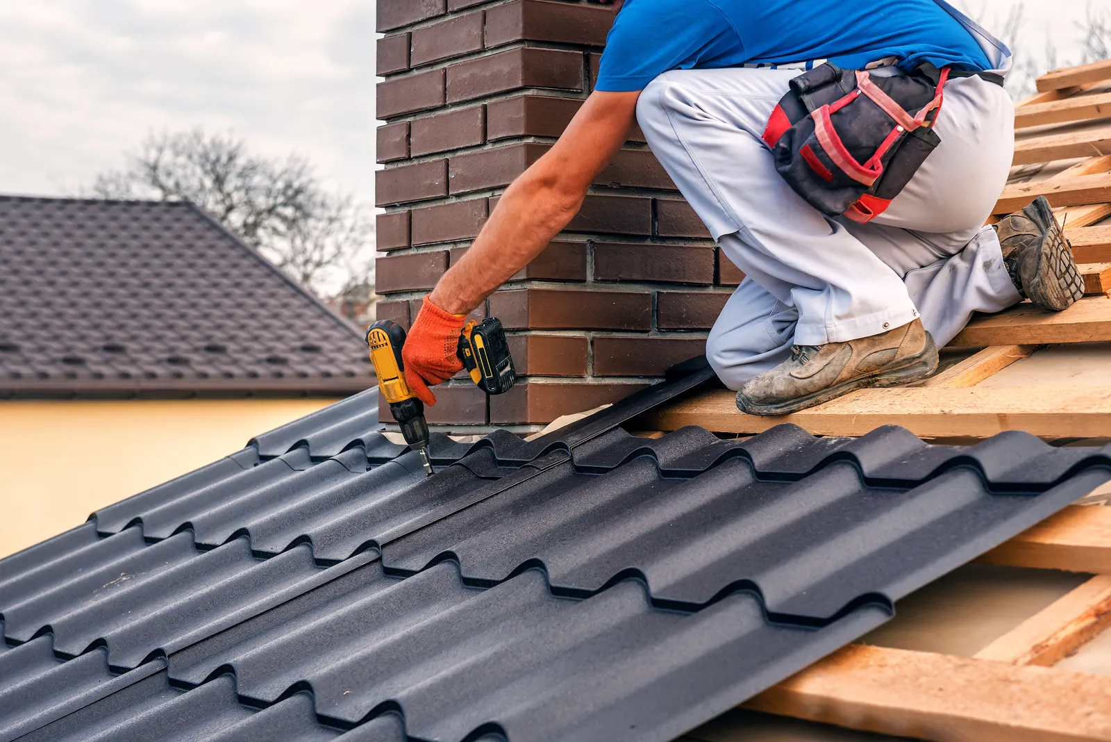 Roofer fitting dark metal tile roof sheets beside a brick chimney