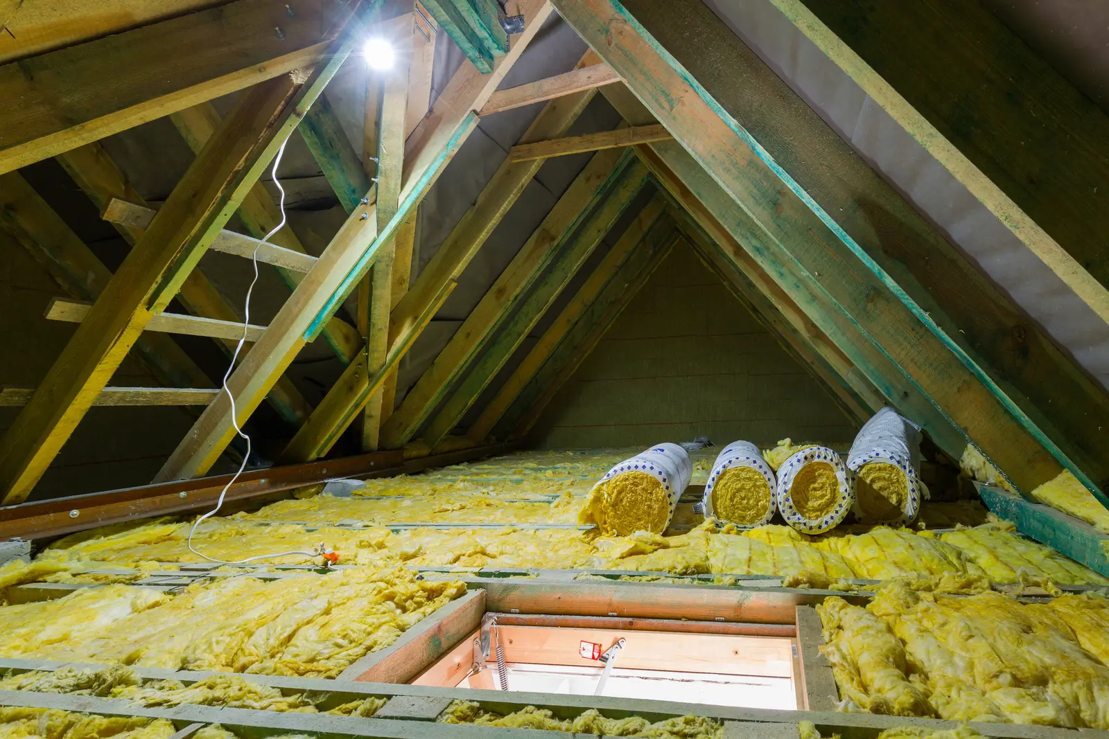 Rolls of mineral wool insulation stacked in an attic roof space above a loft hatch